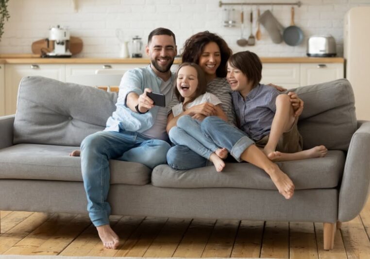 What Are the Signs that I Need a New Furnace? Smiling parents with little kids laughing using smartphone together sitting on couch at home. Happy father holding phone taking selfie with children. Family watching video having fun with cellphone.
